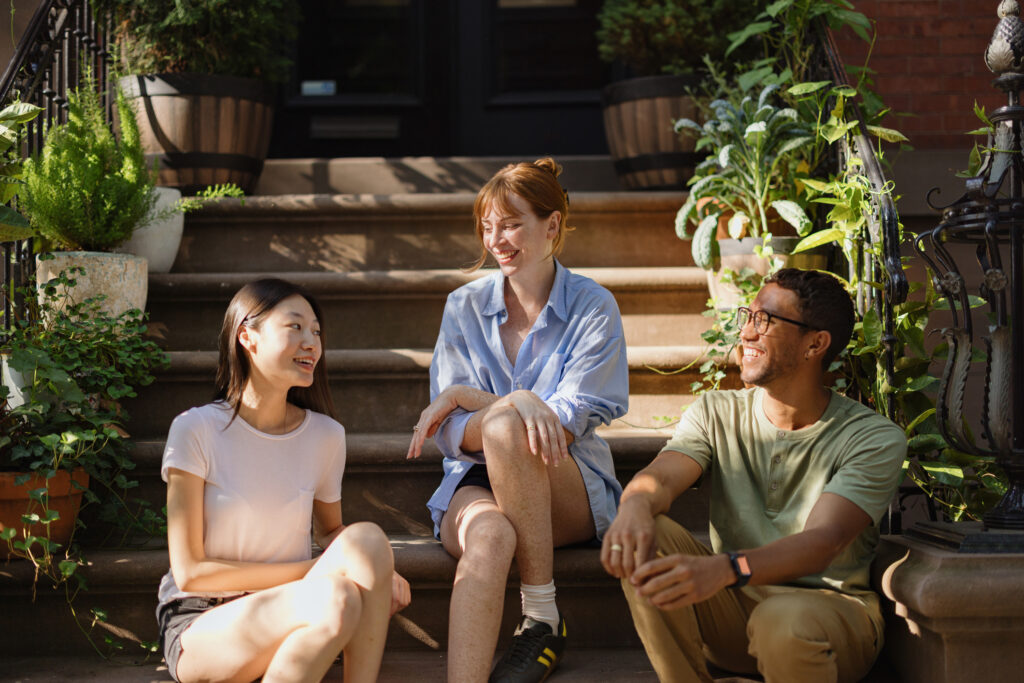 Friends enjoying a conversation while sitting together on stairs surrounded by potted plants. The scene reflects friendship, summer vibes, and the outdoors.